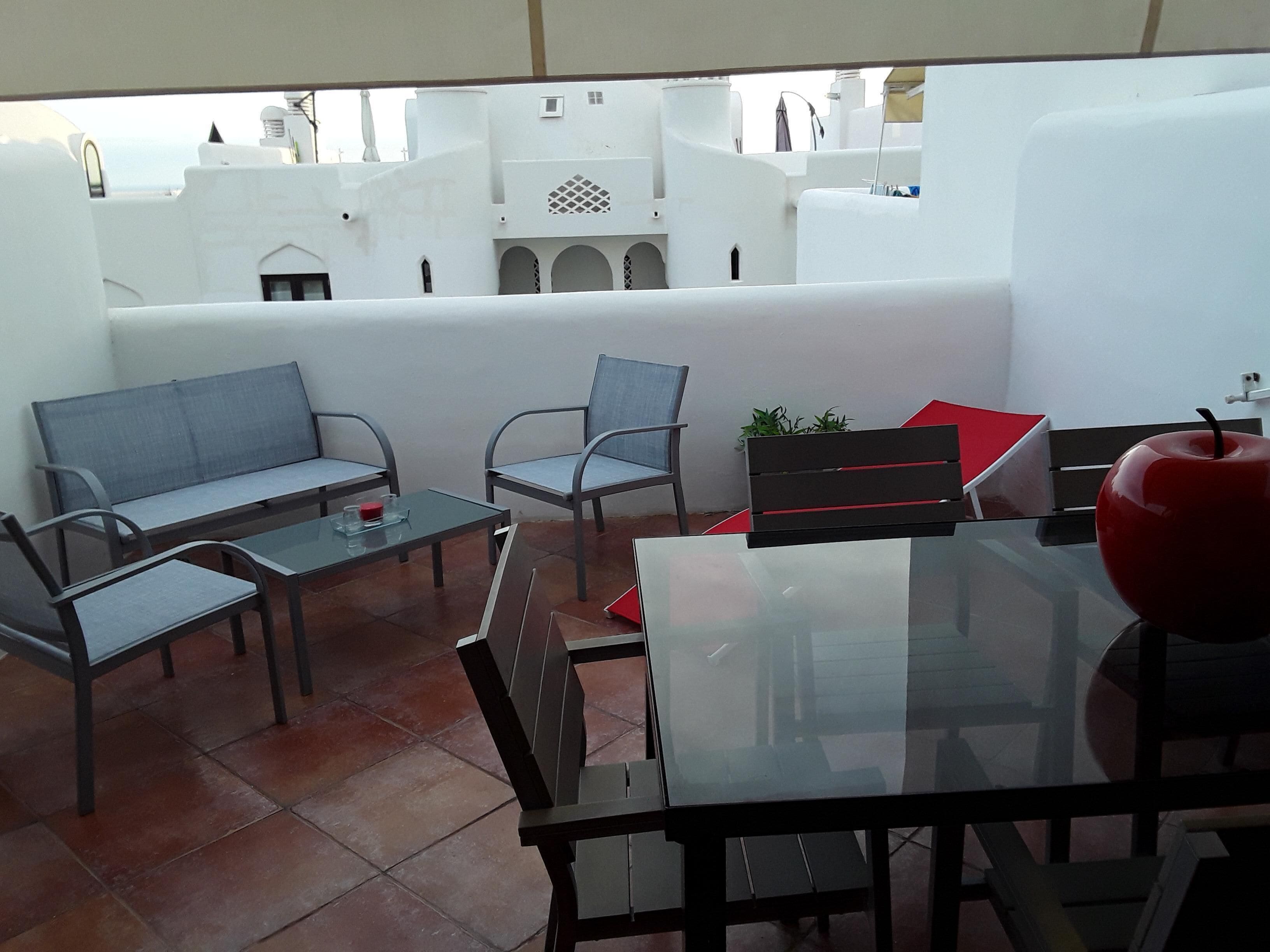 A terrace with a glass table and chairs, a red apple on the table, and a view of white buildings in the background.