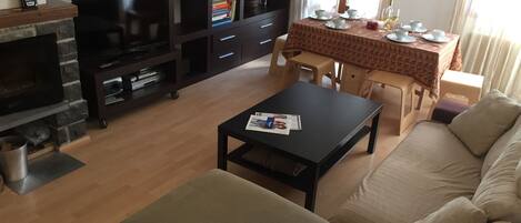 A living room with a sofa, a coffee table, a TV, a fireplace, a bookshelf, and a dining area with a table and chairs.