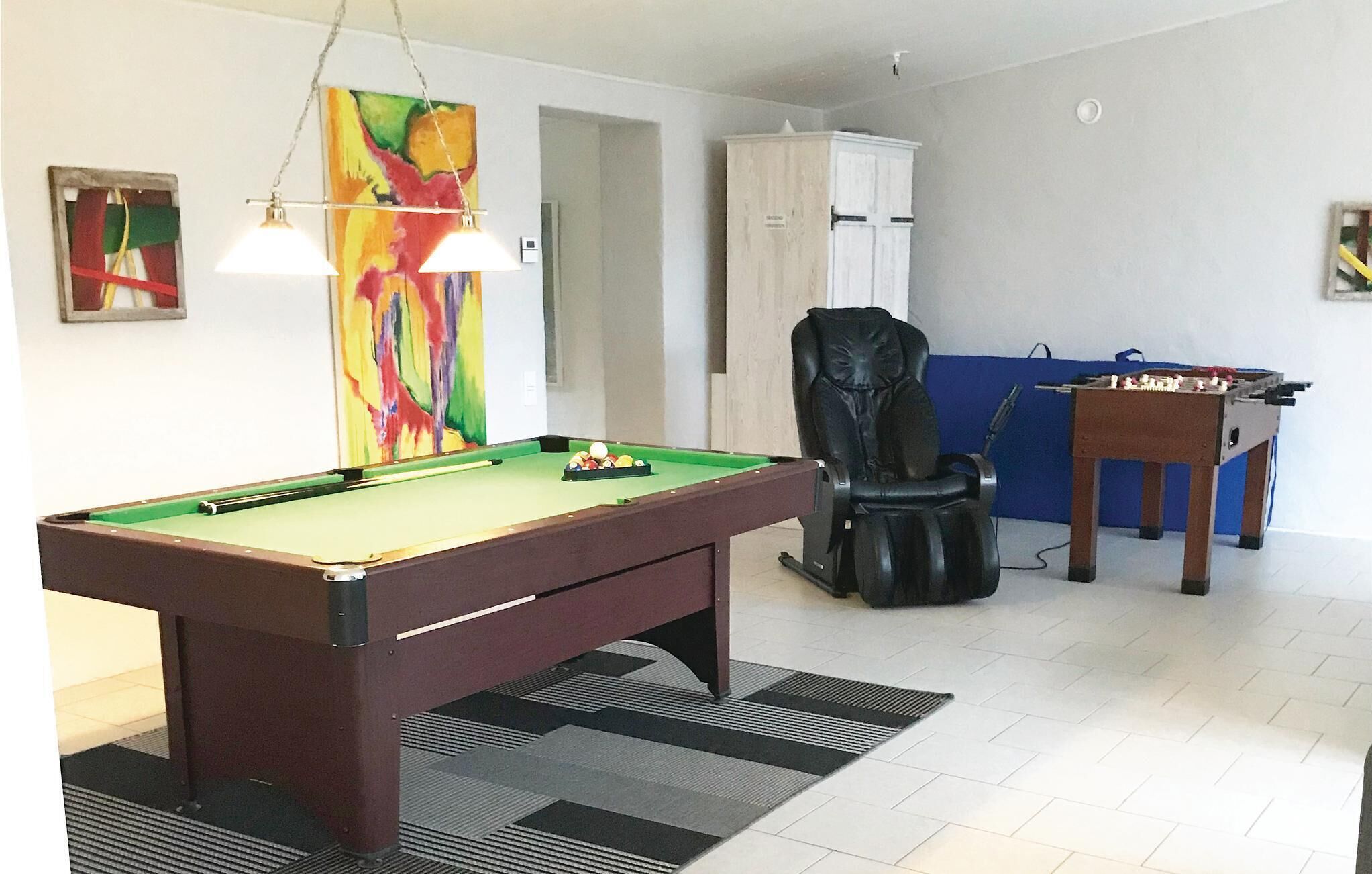 A pool table with a green felt surface, a black leather chair, a foosball table, and a colorful abstract painting on the wall.