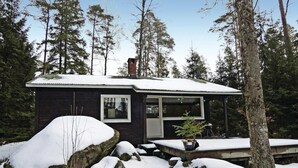 Eine kleine, dunkel gefärbte Holzhütte mit schneebedecktem Dach und Fenstern, umgeben von Bäumen und Steinen.
