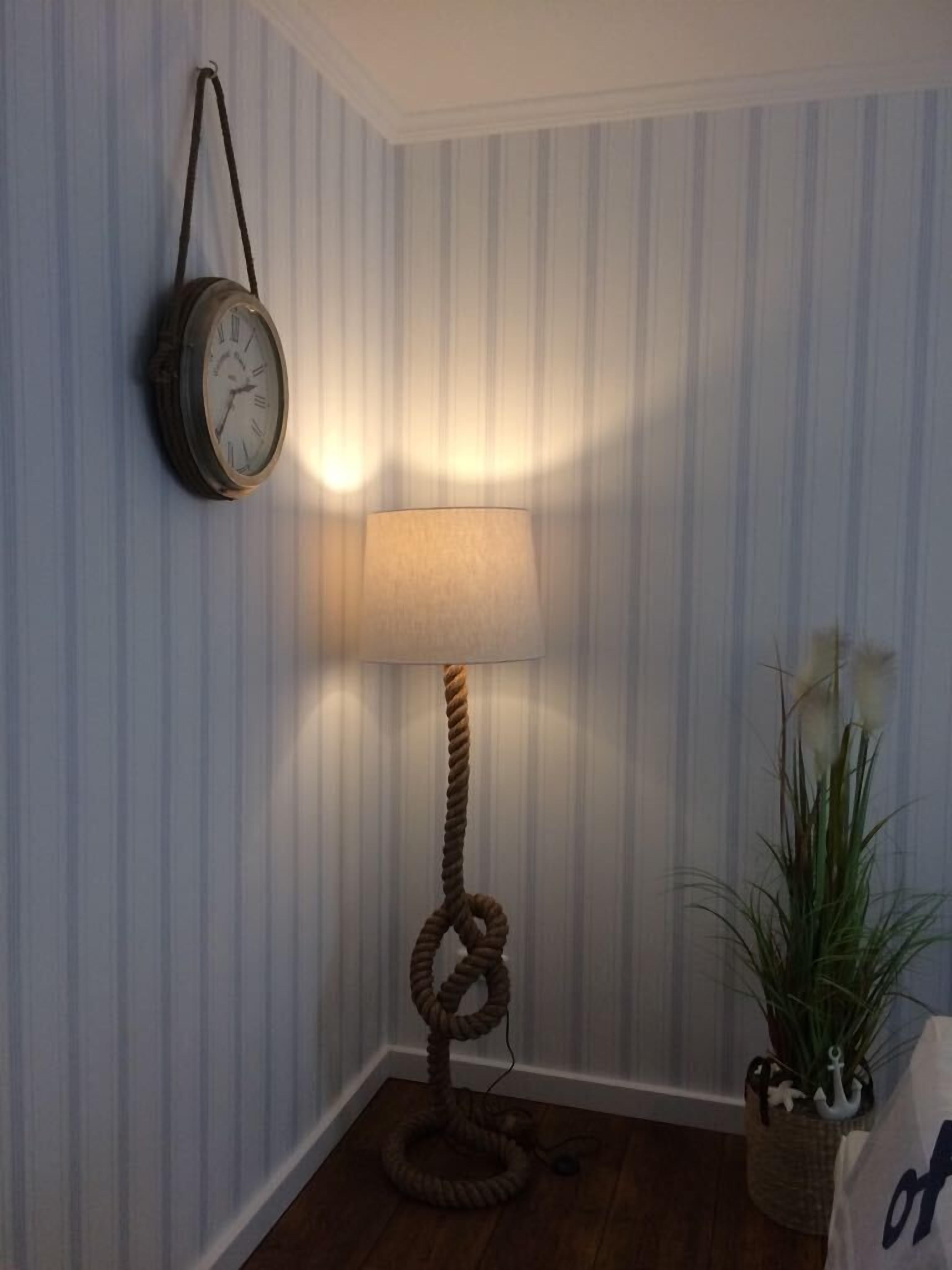 Ein Eckzimmer mit Streifenmuster-Tapete, einer Stehlampe aus Holz, einer Wanduhr und einem Topfpflanzen.