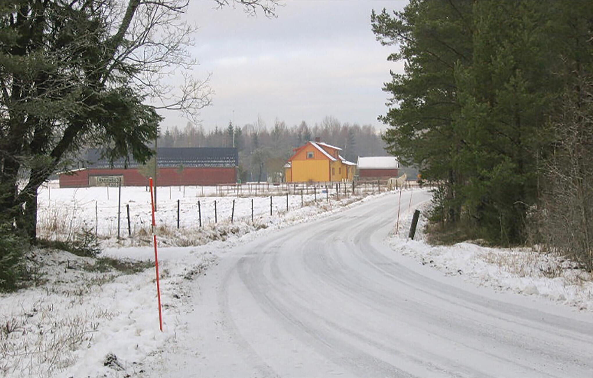 Een landweg met een besneeerd landschap, een gele schuur en een rood gebouw op de achtergrond.
