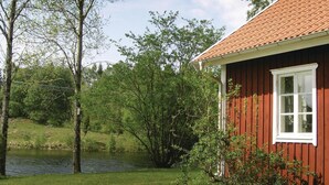 Et rødt træhus med et hvidt vindue, omgivet af grøn vegetation og et vandareal.