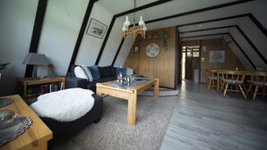 A loft-style living room with a high ceiling, exposed beams, a wooden dining table, and a cozy seating area with a sofa and a coffee table. - Holiday house Fichtelgebirgshaus (Hüttstadl)
