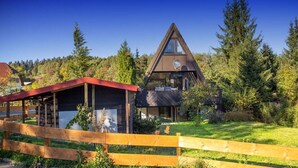 A modern triangular building with a red-roofed structure in the foreground, surrounded by a wooden fence and lush greenery. - Holiday house Fichtelgebirgshaus (Hüttstadl)
