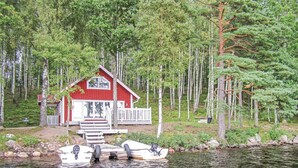 Une cabane à l’extérieur rouge, aux bordures blanches et dotée d’un porche. Elle est située au bord d’un plan d’eau, entourée d’arbres.