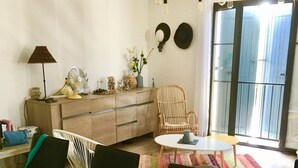 Un comedor con una mesa de madera, sillas negras, una mesita redonda pequeña y un tapete colorido. Hay una silla de mimbre, un ventilador y una lámpara sobre un mueble.