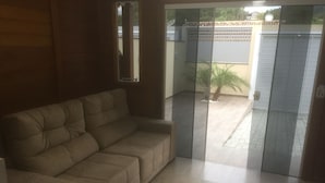 A living room with a beige sofa, a glass door leading outside, and a tiled floor. - Bangalôs do Parque (Penha)