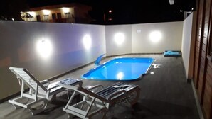 An outdoor pool area with two lounge chairs and a small table, surrounded by walls with lights. - Bangalôs do Parque (Penha)