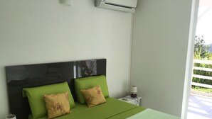 A neatly made bed with green bedding and two decorative pillows, a bedside table with a lamp, and a view of greenery outside.