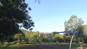 Ein Spielplatz mit Schaukeln und einer Kette, umgeben von Bäumen und blauem Himmel.