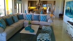 A spacious living room with a beige sectional sofa, a coffee table, and patterned ottomans. A kitchen is visible in the background with a dining area. - The Hacienda Beach Club & Residence 3 bedrm./4 bathroom. (Cabo San Lucas)