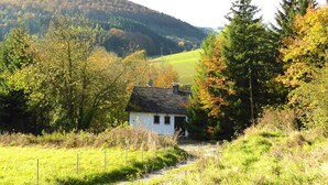 Une maison blanche avec un toit noir, entourée d’arbres et d’un champ d’herbe, avec des collines en arrière-plan.