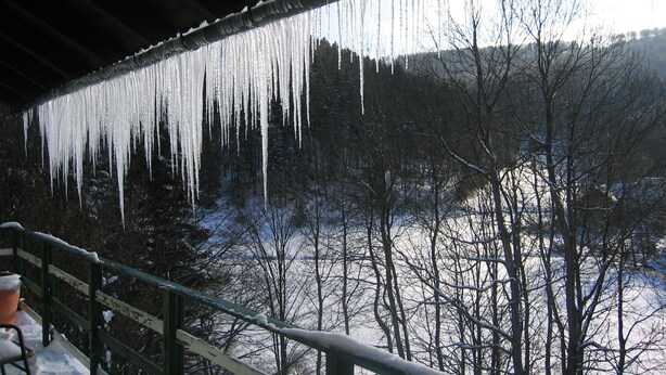 Des glaçons pendants du toit, un paysage recouvert de neige et des arbres sans feuilles.