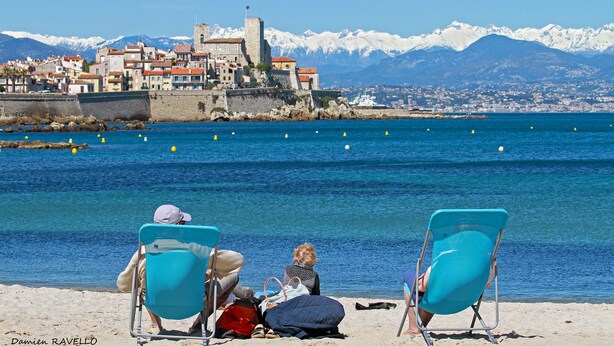 Zwei Strandstühle mit Blick auf eine Küstenstadt, im Hintergrund ein historisches Gebäude und schneebedeckte Berge.