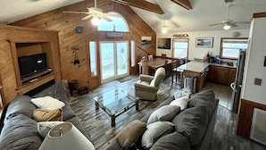 A spacious living room with a large sofa, a coffee table, and a television. There is a dining area with a table and chairs, and a kitchen area with a refrigerator and sink.