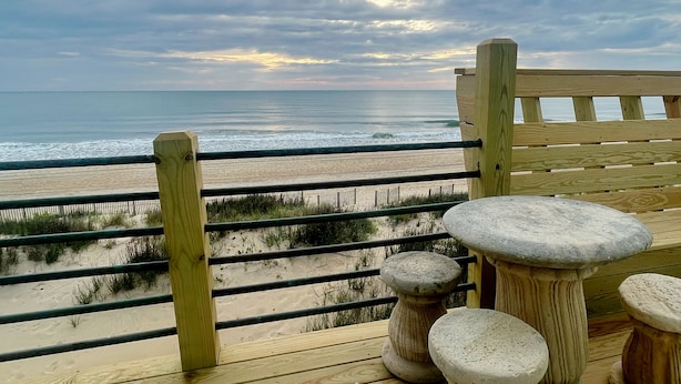 A wooden deck with a bench and stone stools overlooking a beach with sand dunes and the ocean.