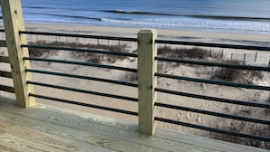 A wooden deck with metal railings overlooking a beach with sand and sparse vegetation.