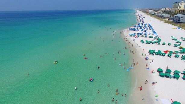 Ein Strand mit klarem türkisblauem Wasser und weißem Sand, gesäumt von Reihen grüner Sonnenschirme.