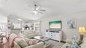 A modern living room with a sofa, coffee table, and a TV. There is a kitchen in the background with white cabinets and a microwave.