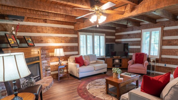 A log cabin interior with a fireplace, wooden floors, a ceiling fan, and a cozy seating area. - Cozy & Chic Rustic Retreat-Luxury Log Home Near Downtown, Opry and Much More (Nashville)