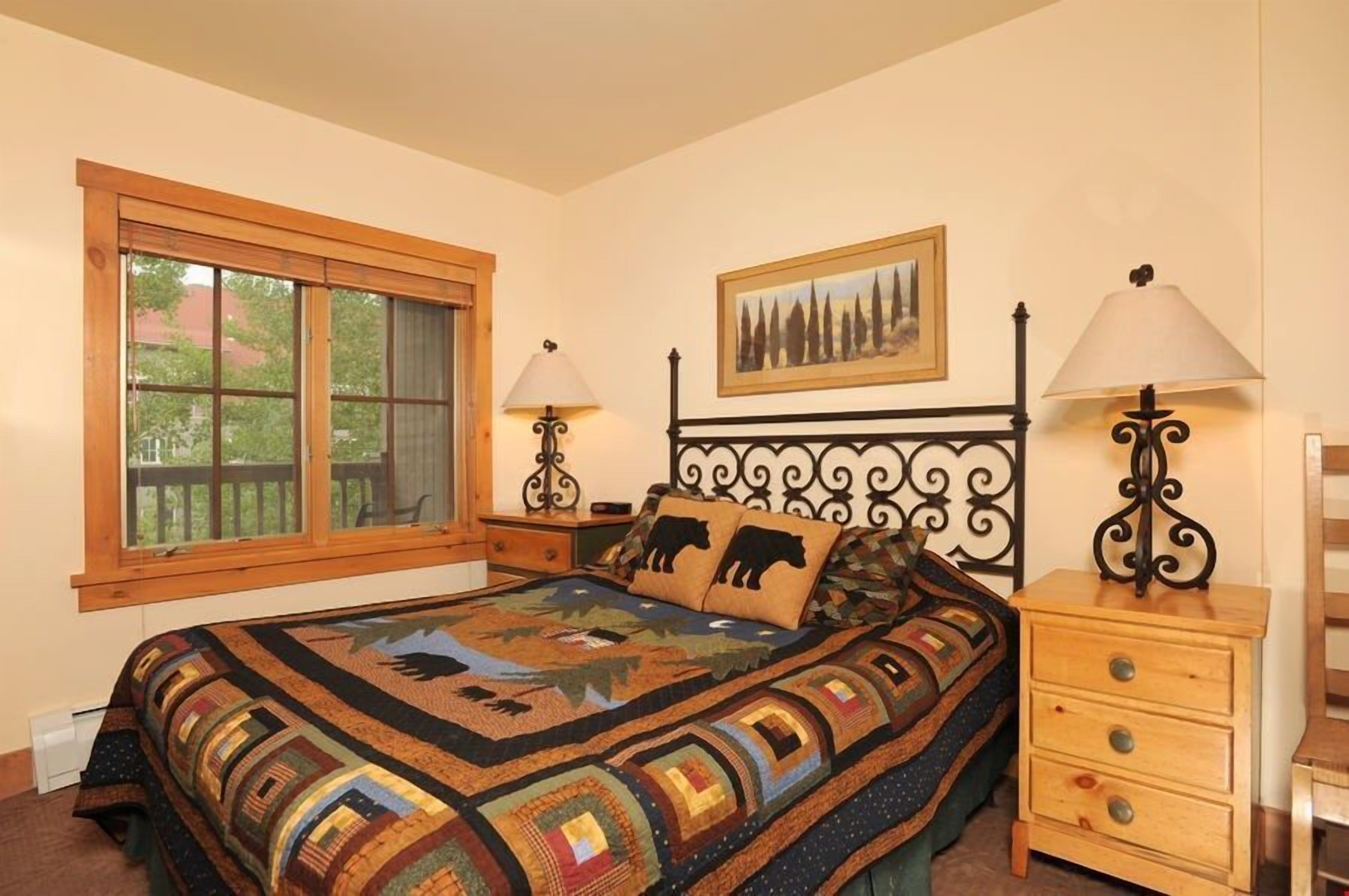 A bedroom with a large window, a bed with a quilt, two bedside tables with lamps, and a painting on the wall.