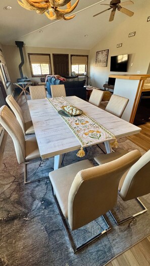 Un comedor con una mesa larga, sillas acolchadas, una araña y una cocina al fondo.