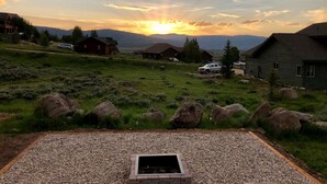 A gravel patio with a fire pit, surrounded by a grassy field with large rocks, and houses in the background under a sunset sky. - 1 Mile to ski Area / Amazing Views / Hot Tub - Five Star Reviews (Granby)
