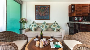 A cozy living area with wicker furniture, a wooden coffee table with candles and a book, a sofa with patterned cushions, and a kitchen area with wooden cabinets.