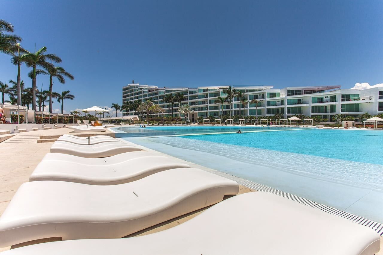 A poolside area with lounge chairs, a clear blue pool, palm trees, and a modern hotel building in the background.