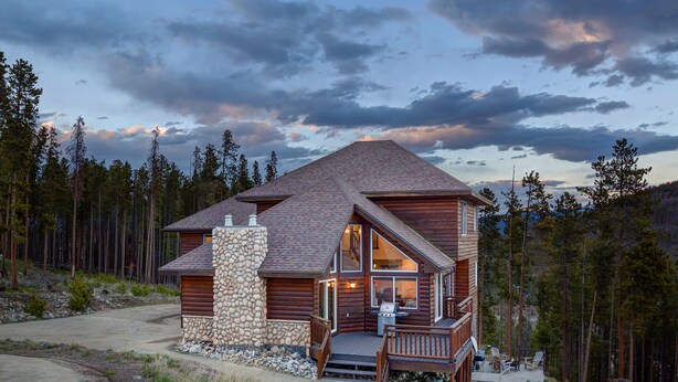 Una cabaña de troncos con chimenea de piedra, ventanas grandes y una terraza con mobiliario exterior.