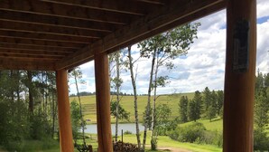 Un espacio exterior cubierto con pilares de hormigón, una leñera y vistas a un lago y un paisaje verde.