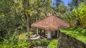 A thatched-roof hut with a glass door, surrounded by lush greenery and a stone wall with carvings. - Elegant, Intimate, Luxury Contemporary Balinese Villa  In a Traditional Village  (Kemenuh, Sukawati,)