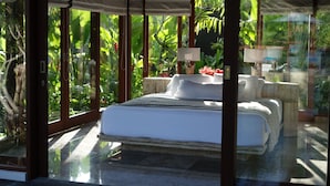 A bedroom with a large bed, a nightstand, a lamp, and a view of lush greenery through large windows. - Elegant, Intimate, Luxury Contemporary Balinese Villa  In a Traditional Village  (Kemenuh, Sukawati,)