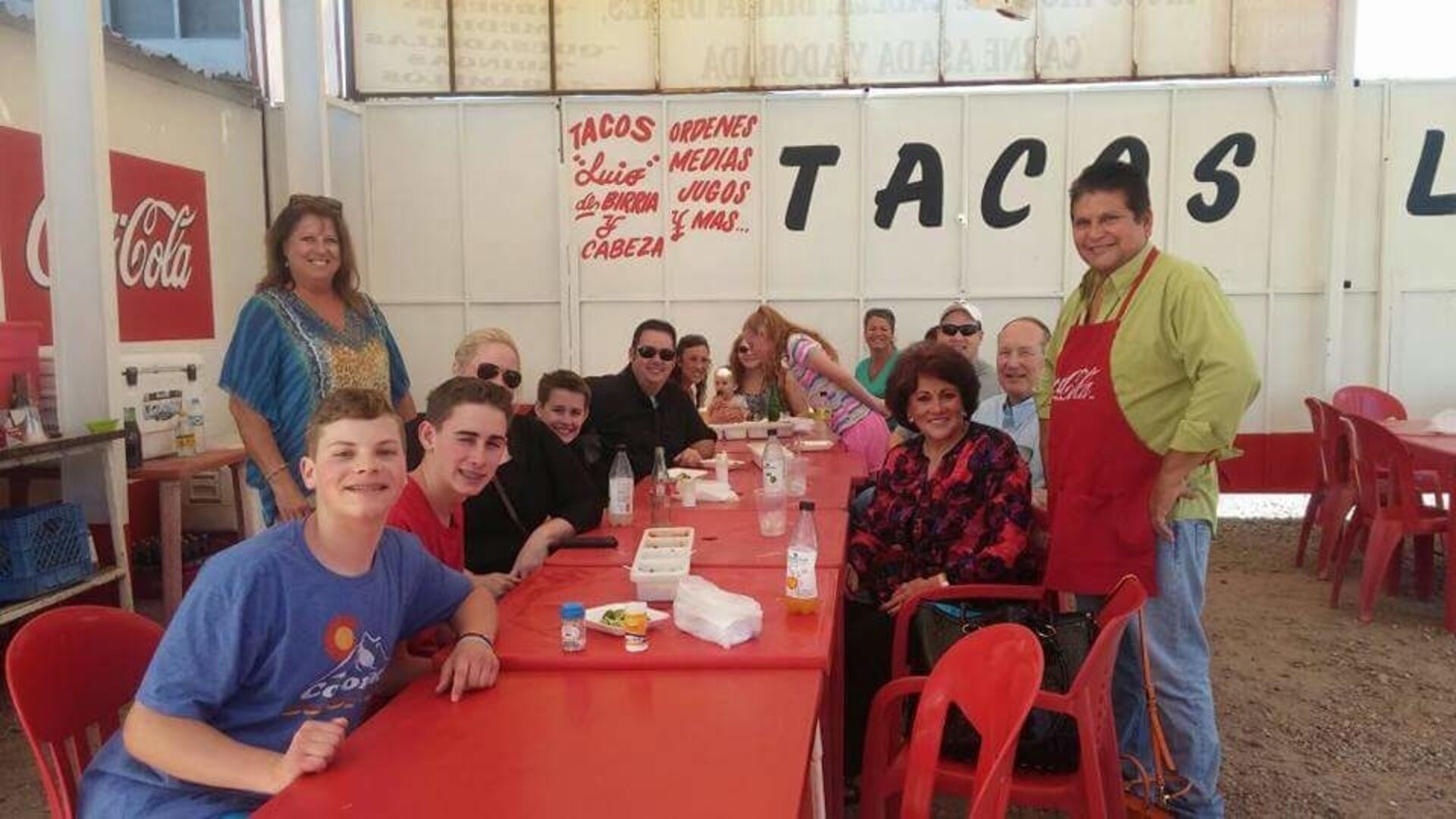 Um grupo de pessoas sentadas à mesa vermelha, com comidas e bebidas, em um ambiente descontraído para refeições, com um letreiro da Coca-Cola ao fundo.