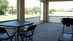 A covered patio area with a table and chairs, a grill, and a view of a grassy area with trees and a fence. - beautiful homeaway , 2815 sw 29th pl (Cape Coral)