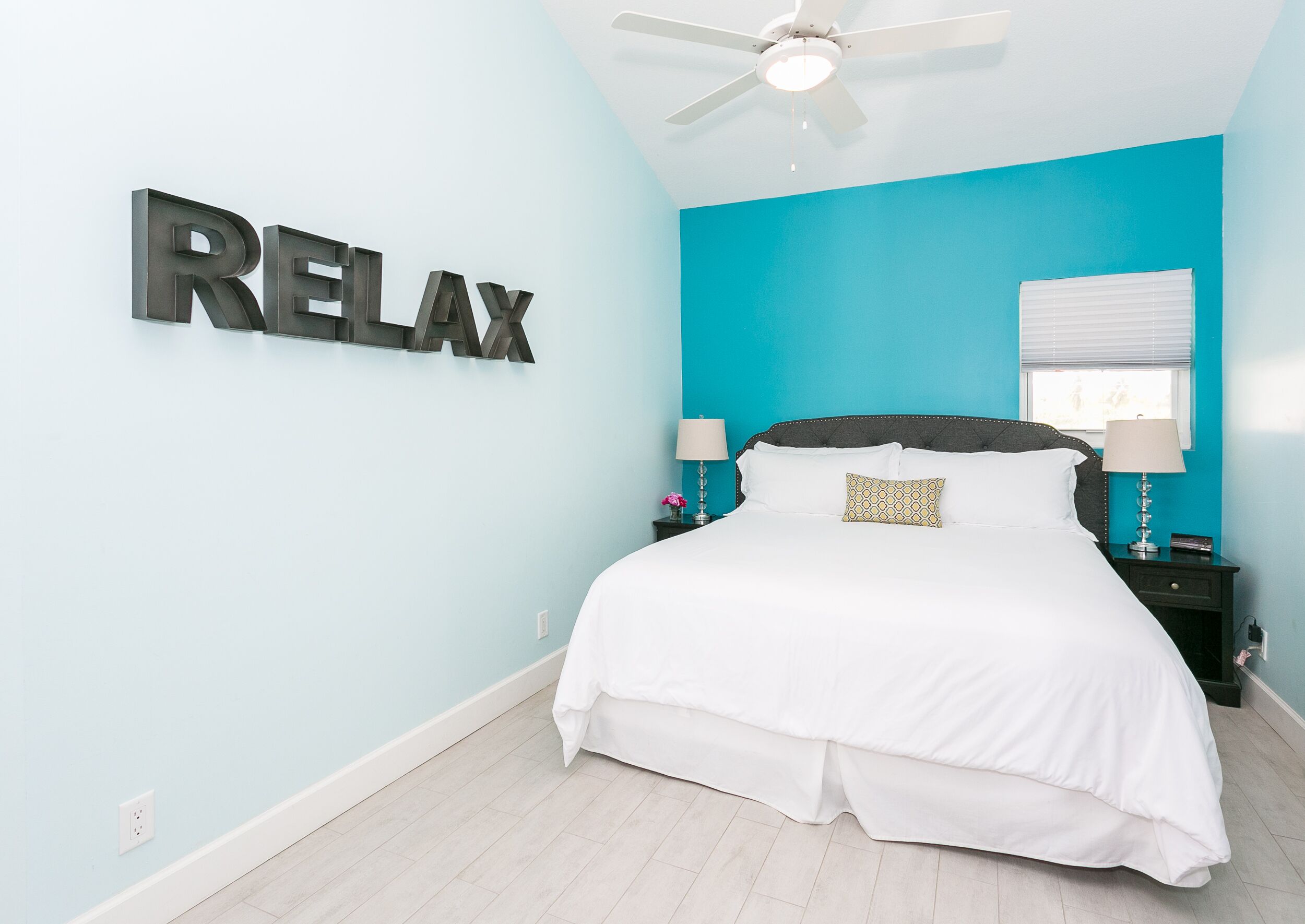 Un dormitorio con cama, ventilador de techo, un cartel en la pared que dice "RELAX" y ventana con persianas.