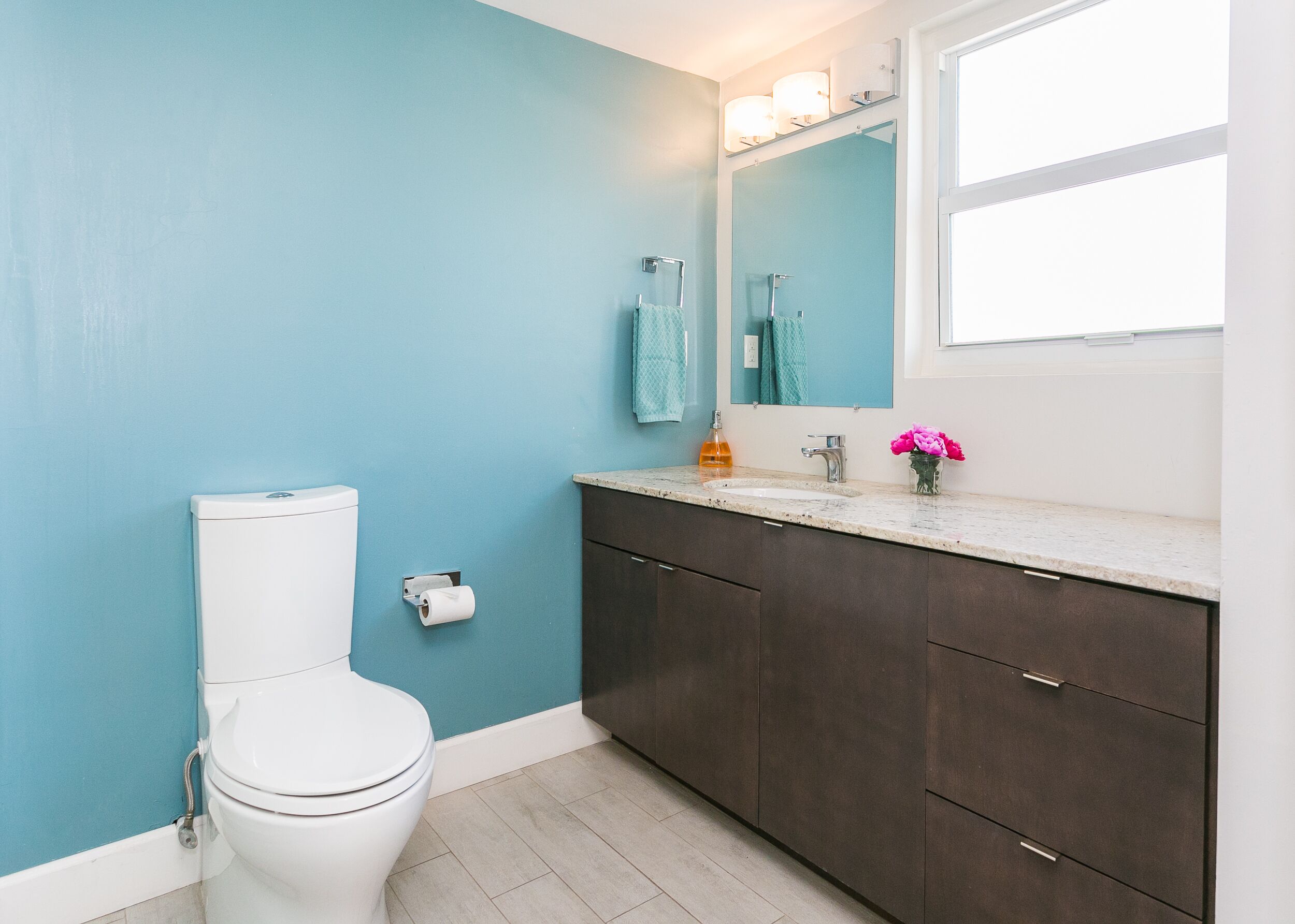 A bathroom with a white toilet, a sink with a marble countertop, dark cabinets, a mirror, and a window.