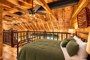 A wooden interior with a sloped ceiling, a bed with green bedding, and a balcony railing.