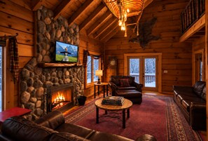A cozy log cabin interior with a stone fireplace, leather sofas, a television mounted on the wall, and a large window overlooking a snowy landscape.