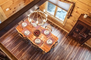 Een houten eettafel met een kroonluchter, omringd door stoelen, in een blokhut met houten wanden en een raam.