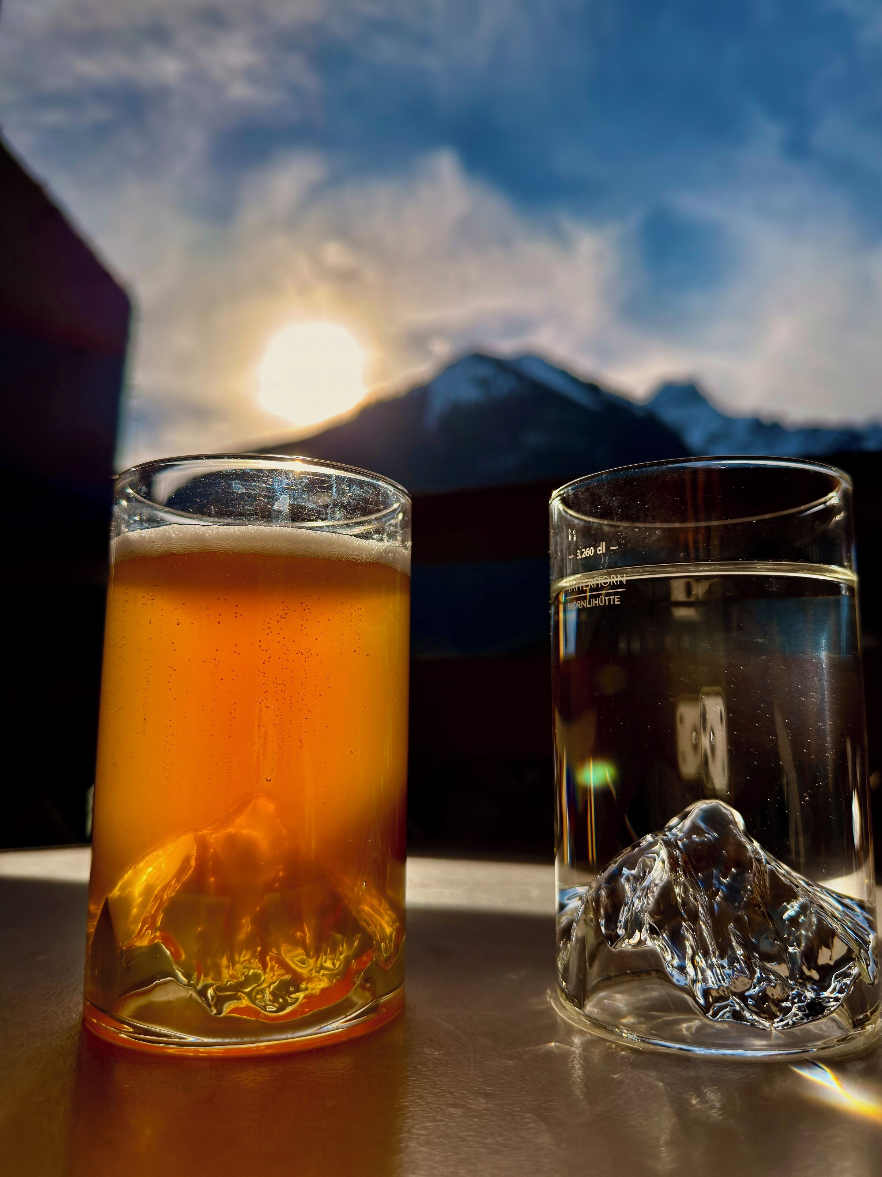 Two glasses of liquid, one with ice, on a table with a mountain view in the background.