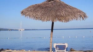 Ein Strand mit einem Strohdachschirm und einem Stuhl, ruhiges Meer und Boote in der Ferne.