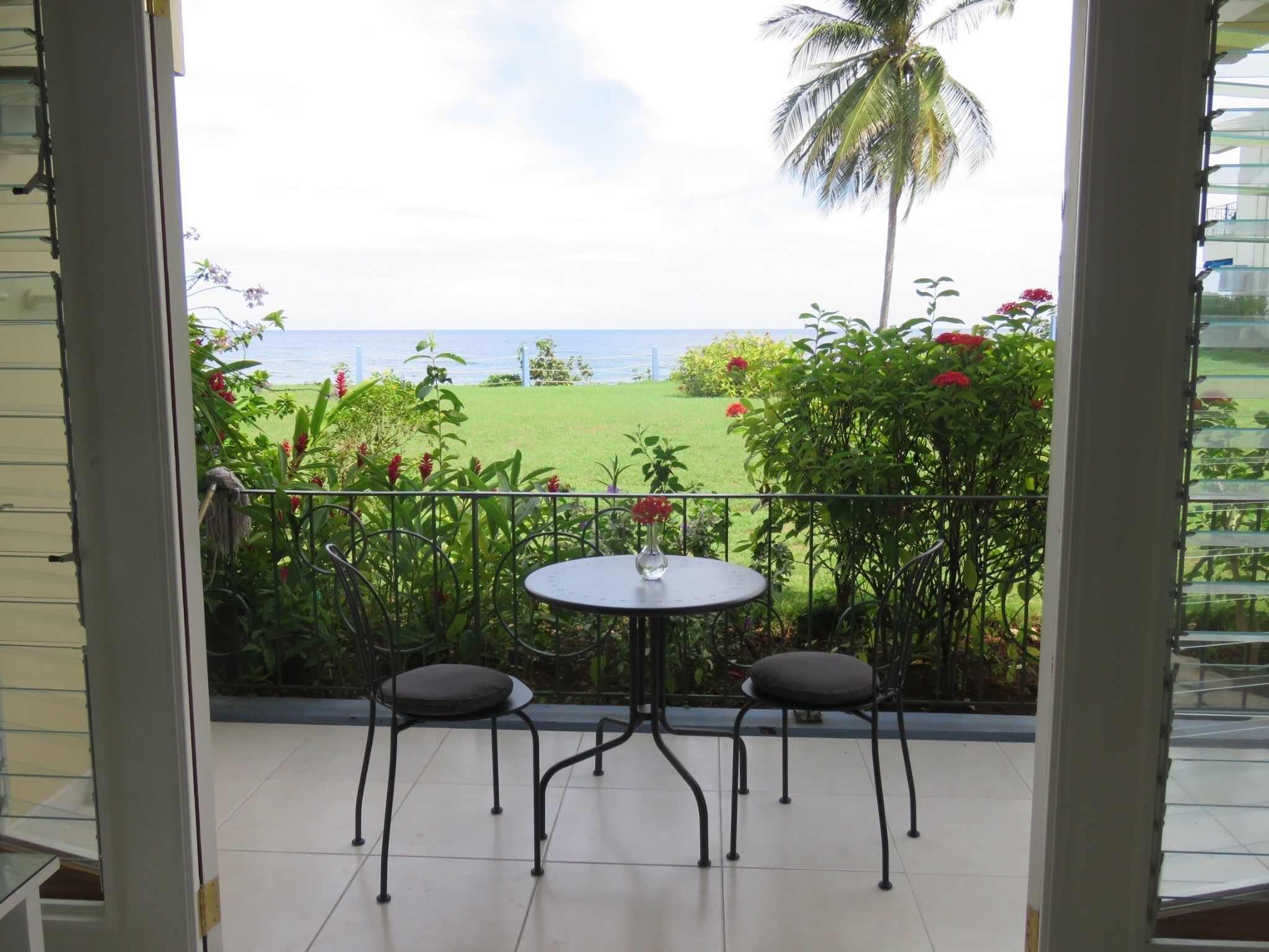 A balcony with a table and two chairs, overlooking a grassy area with a view of the sea and palm trees.