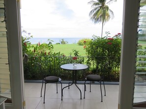 A balcony with a table and two chairs, overlooking a grassy area with a view of the sea and palm trees.