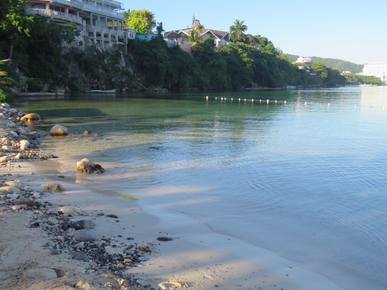 Un río tranquilo con orilla rocosa y edificios al fondo.