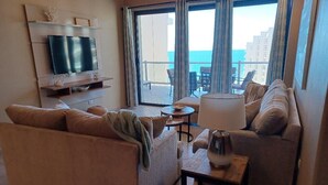 Een woonkamer met een bank, een salontafel en een aan de muur bevestigde televisie. Er is een balkon met uitzicht op zee en gebouwen.