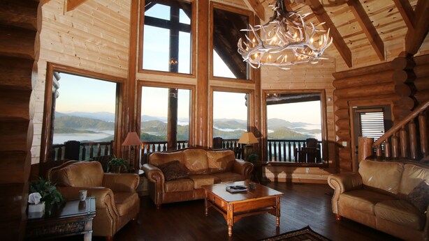 A log cabin interior with leather sofas, a wooden coffee table, and a chandelier. Large windows offer views of mountains. - Majestic Mountaintop Retreat- Breathtaking Views, Luxury Lodge over Pigeon Forge (Sevierville)