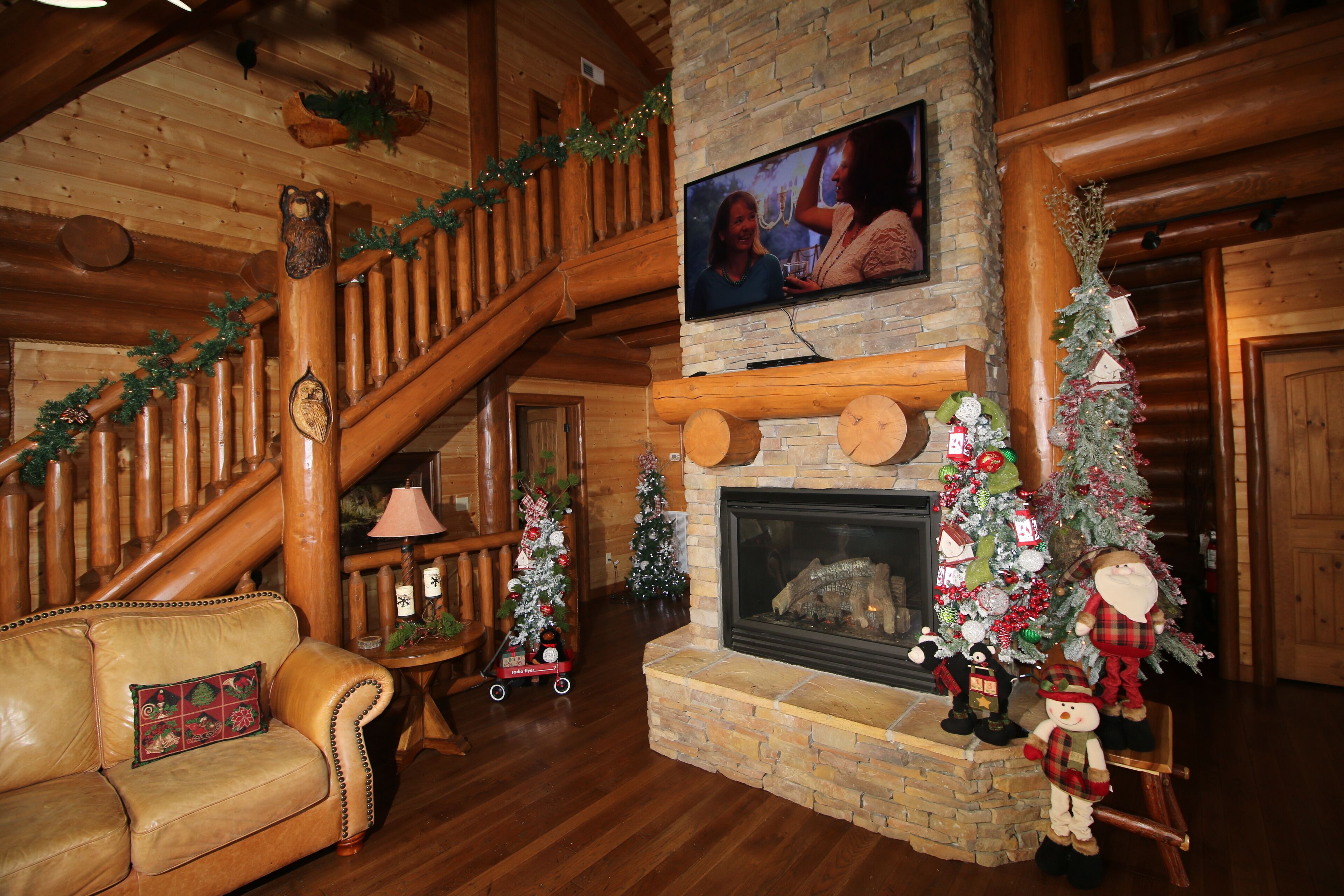 A log cabin interior with a fireplace, Christmas decorations, a leather sofa, and a television mounted on the wall.