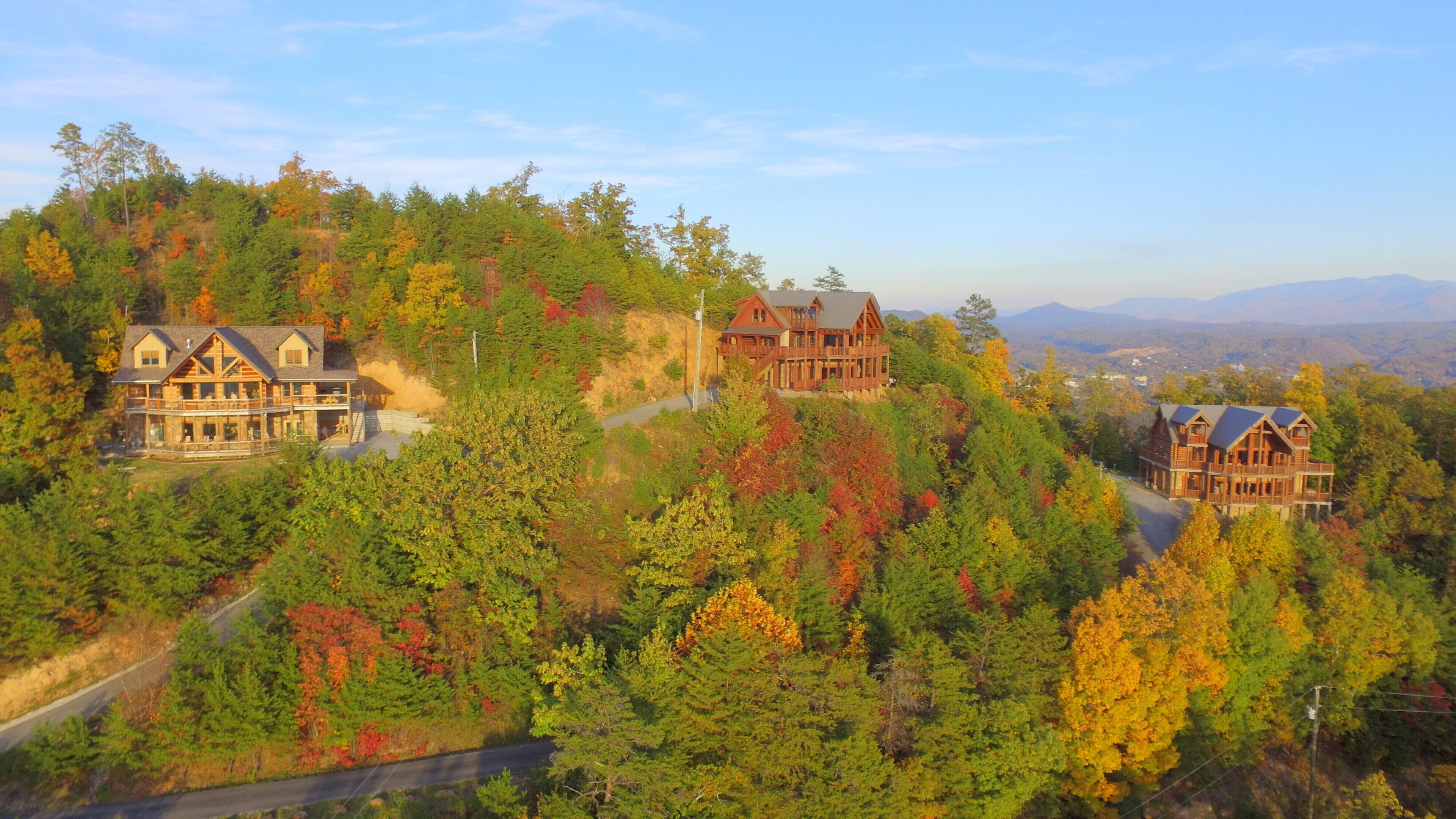 A large, multi-story wooden cabin with a balcony, surrounded by autumn-colored trees and a mountainous backdrop.